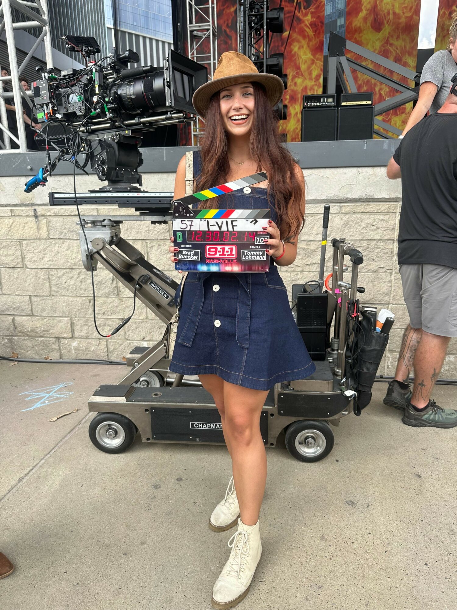 Woman holding a clapperboard on a film set, smiling, wearing a denim skirt and white sneakers.