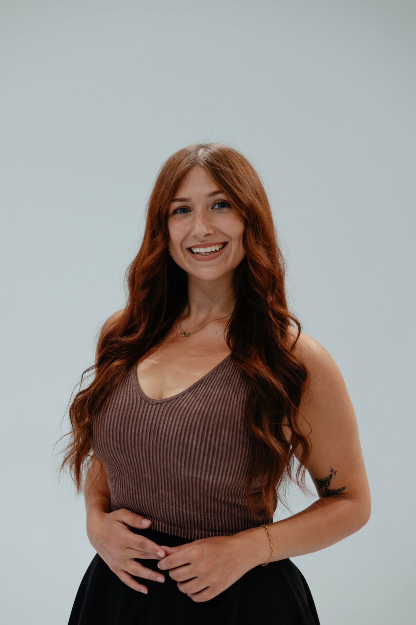 Woman with long wavy red hair smiling, wearing a sleeveless brown top and black skirt, standing against a light background.