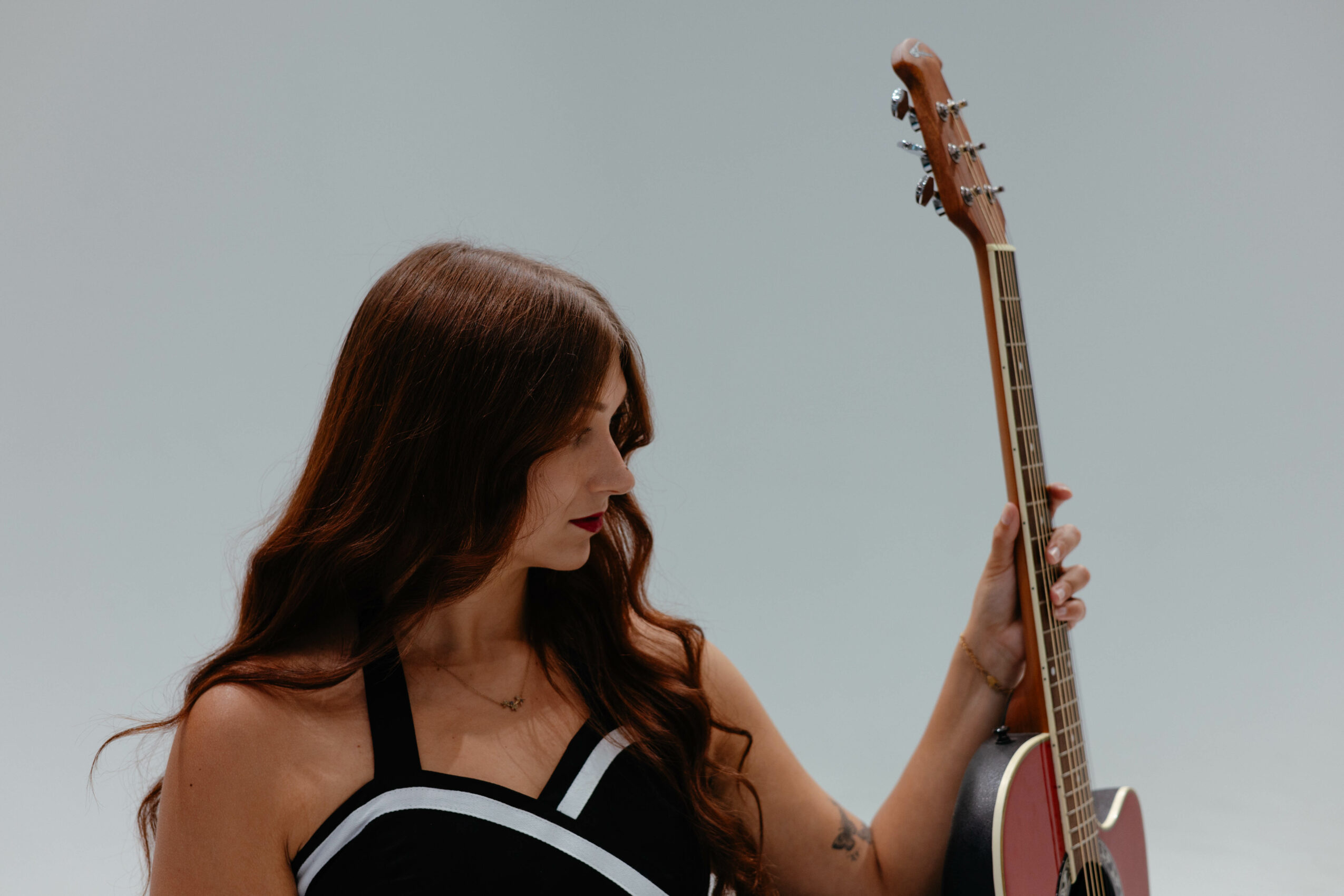 Woman with long brown hair holding an acoustic guitar, wearing a black and white top, against a plain background.