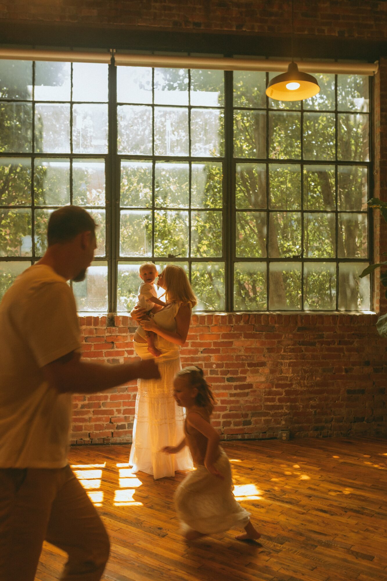 People in a room with large window, sunlight, brick wall, and wooden floor, with a person holding a child and a girl kneeling.