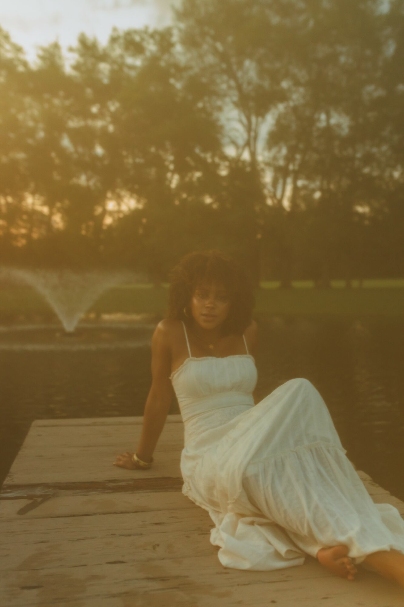 Woman in white dress sitting on wooden dock near water with trees and sunset in background