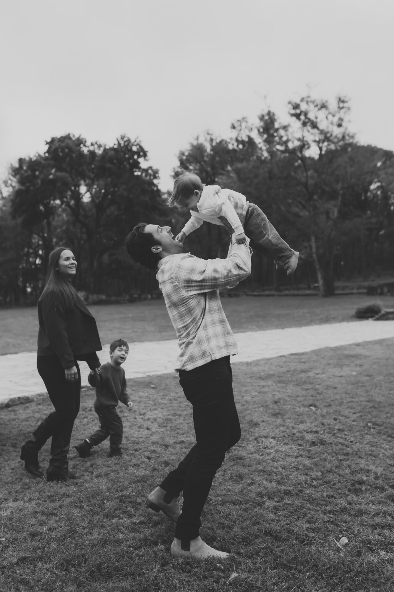 Group of people outdoors, with a person lifting a child, others walking nearby, trees in background, black and white photo.