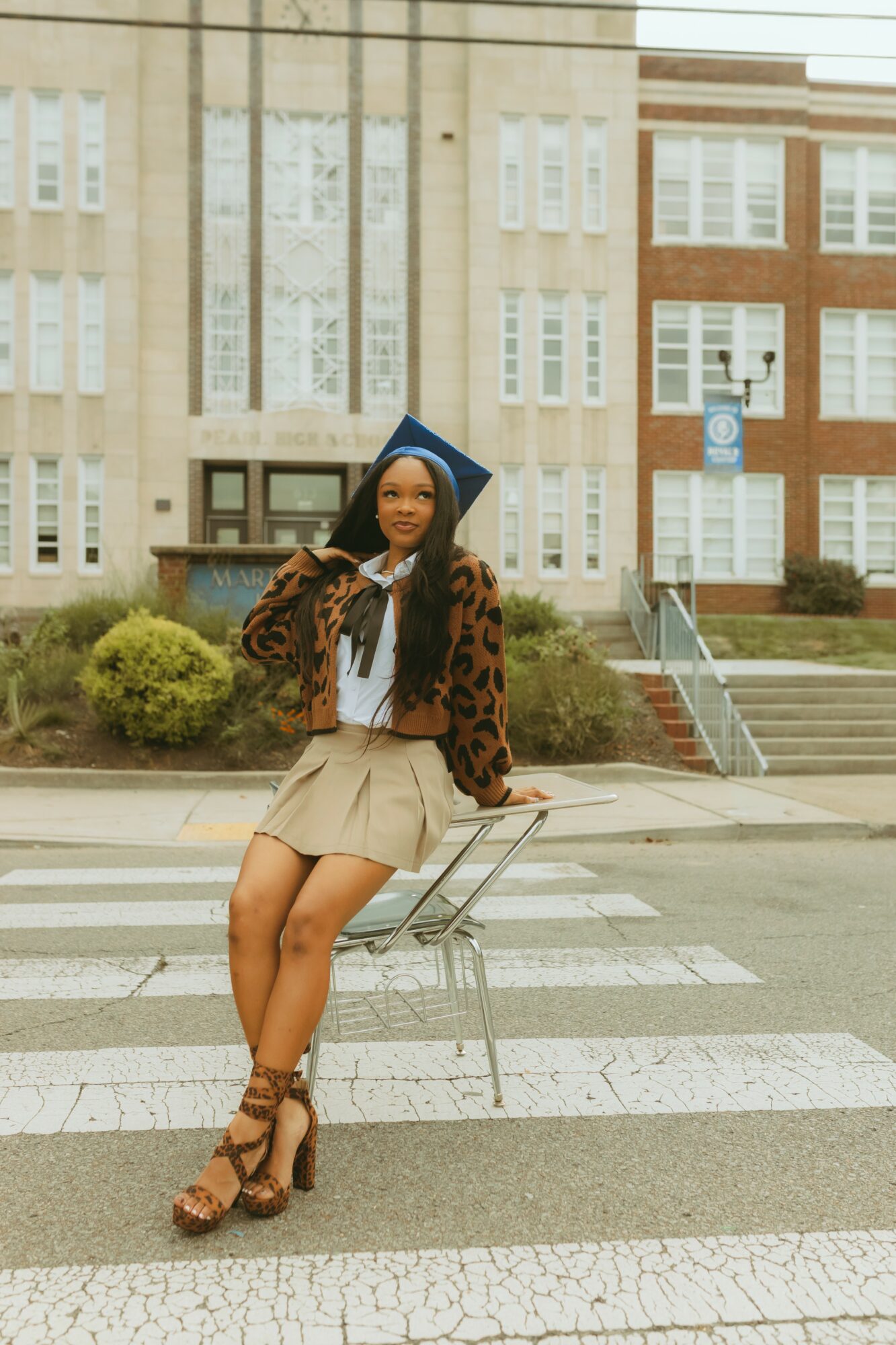 Young woman in graduation cap and gown sitting on a chair outdoors, with buildings in the background.