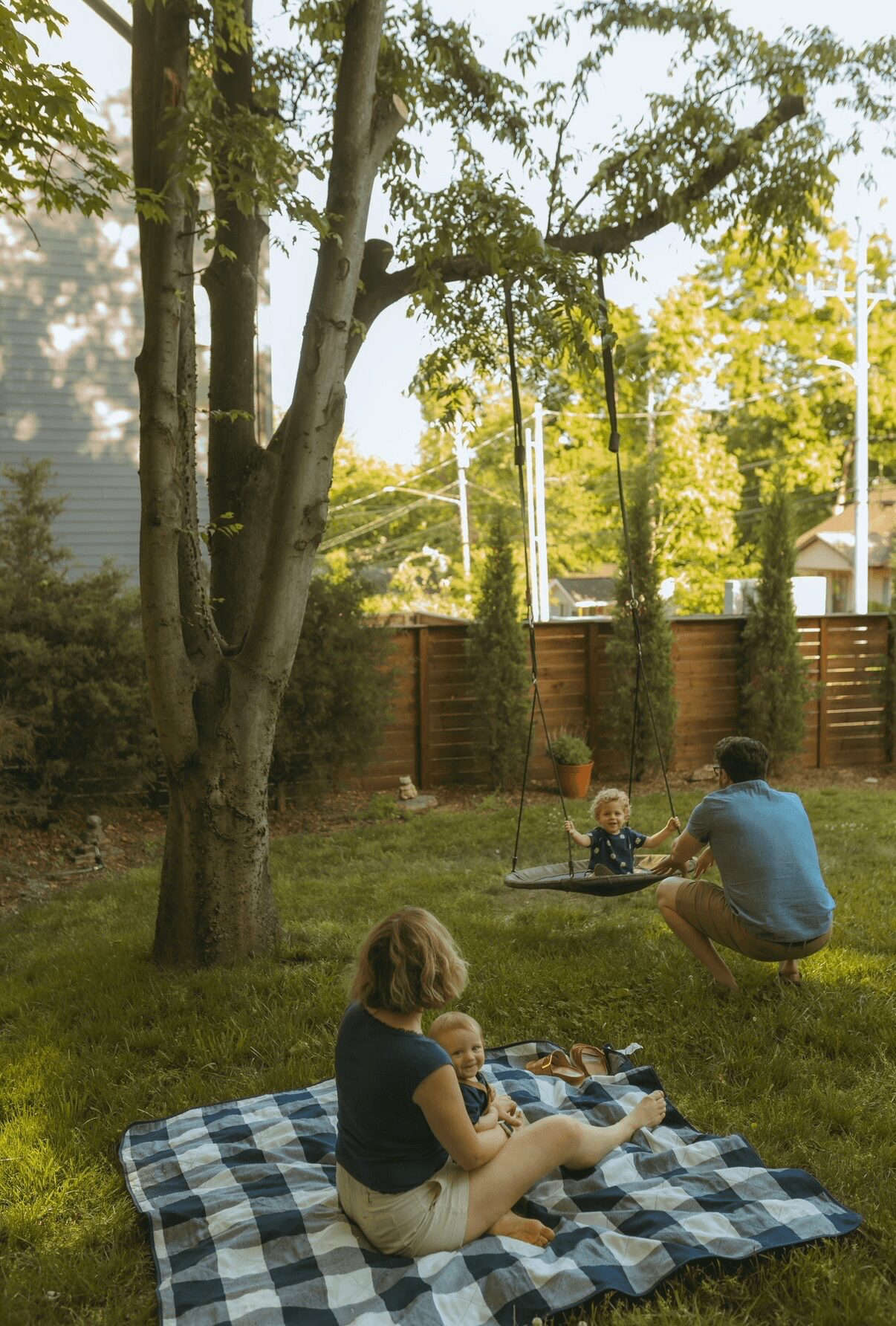 Child sitting on a checkered blanket in a backyard, with a person on a swing nearby, trees and a fence in the background.