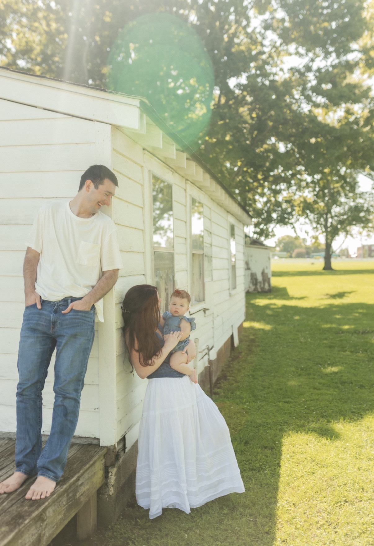 Three people outdoors near a white building, with trees and grass in the background, sunlight shining through leaves.