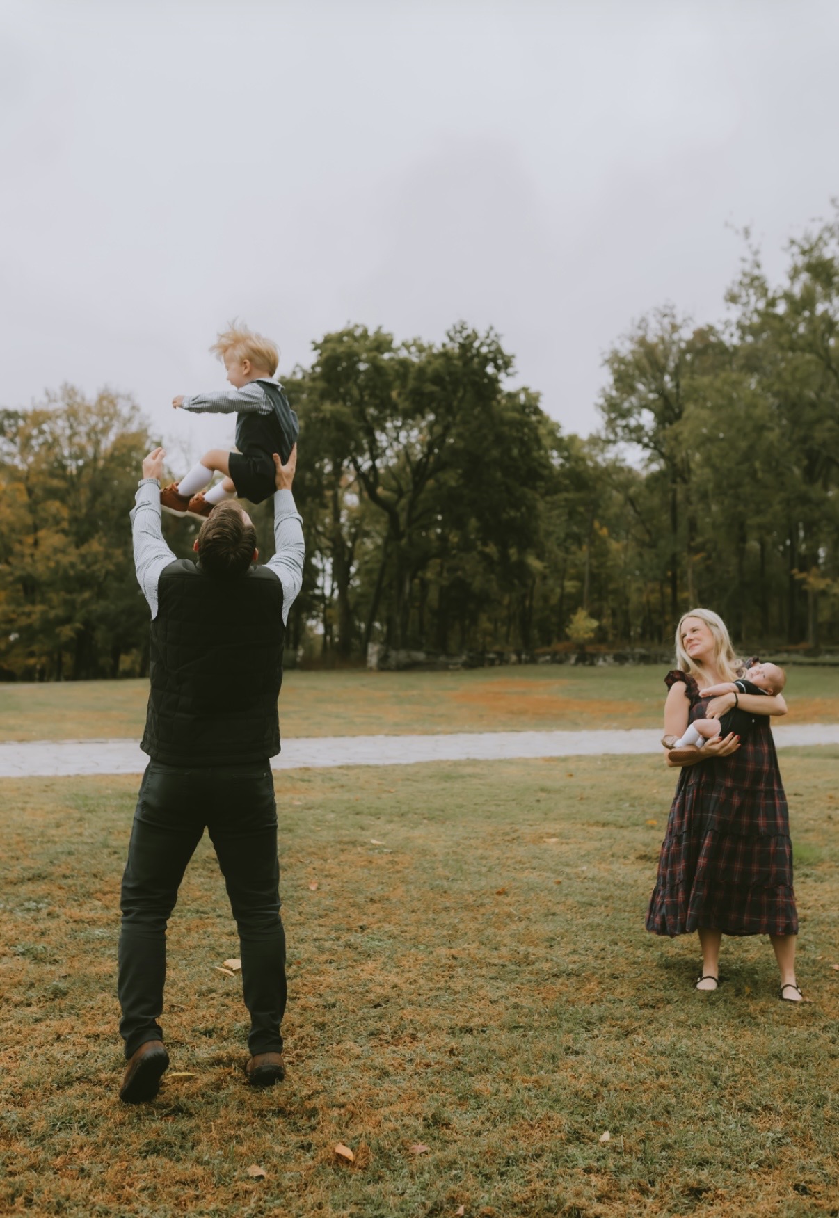 Person holding a child high in the air outdoors, with another person standing nearby holding a dog, trees in background.
