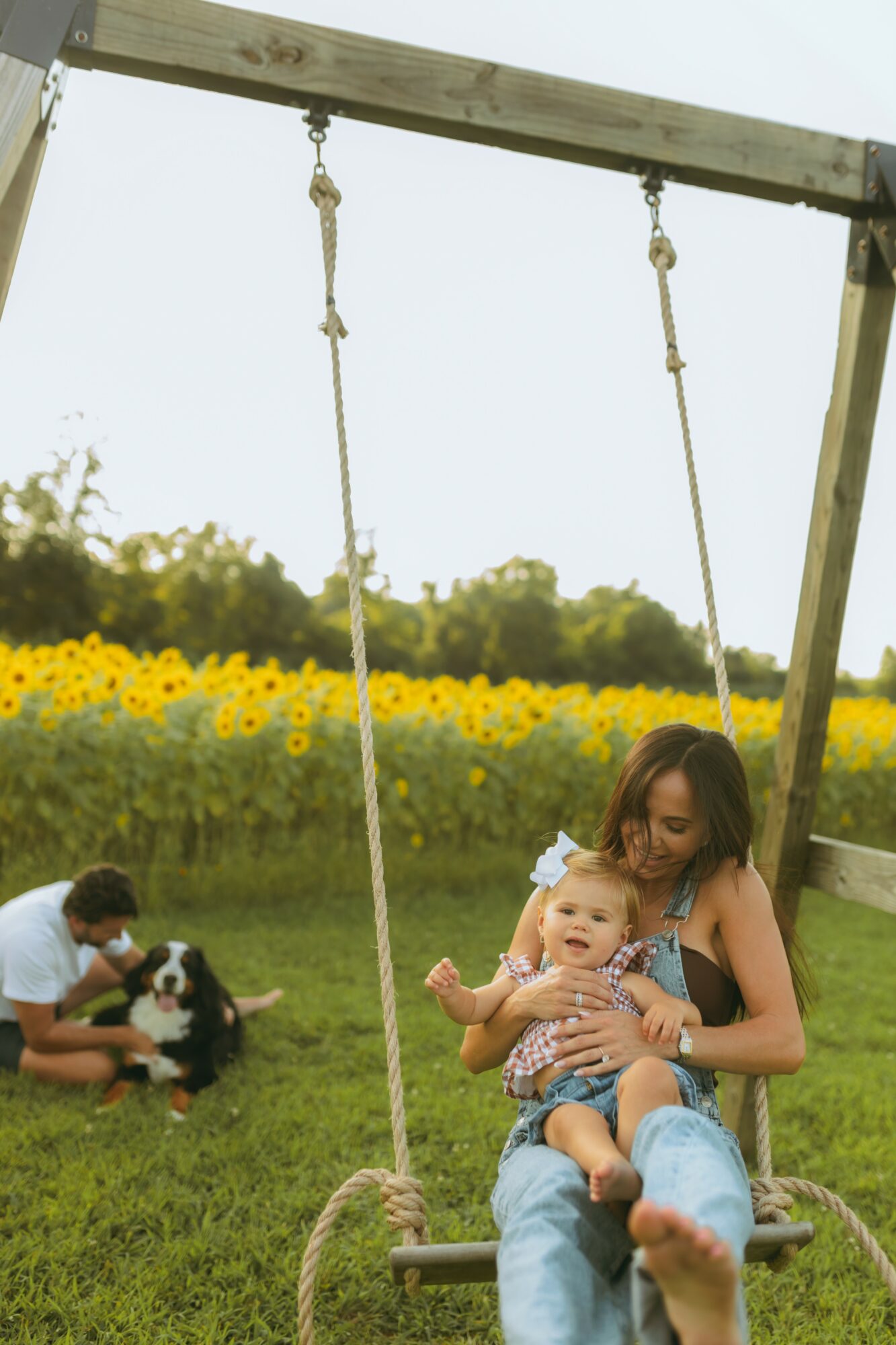 Woman sitting on a swing holding a baby, with a sunflower field and children playing nearby outdoors.