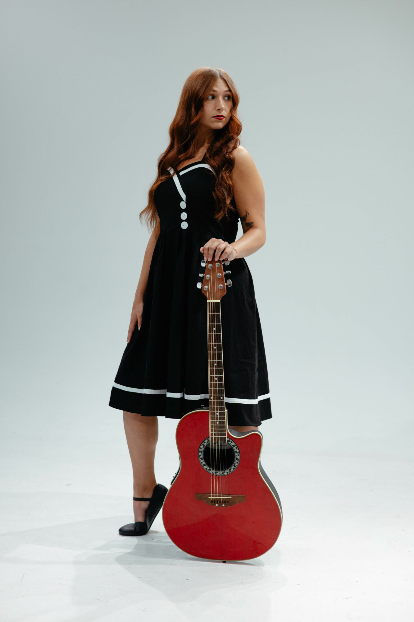 Young woman with long wavy hair in a black dress holding a red acoustic guitar, standing against a plain light background.