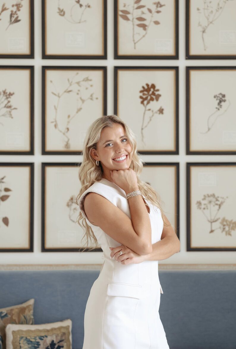 Woman with long blonde hair smiling, standing in front of framed botanical illustrations on a wall.
