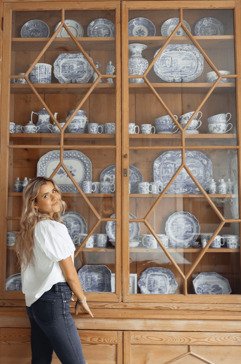 Young woman with blonde hair in a white top and jeans stands in front of a wooden display cabinet filled with blue and white china dishes.