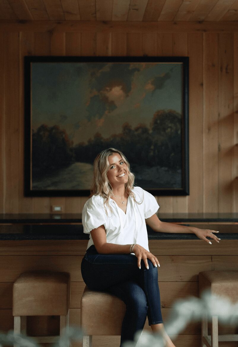 Woman sitting on a wooden bench in front of a landscape painting, smiling and looking upward, with a wooden wall background.
