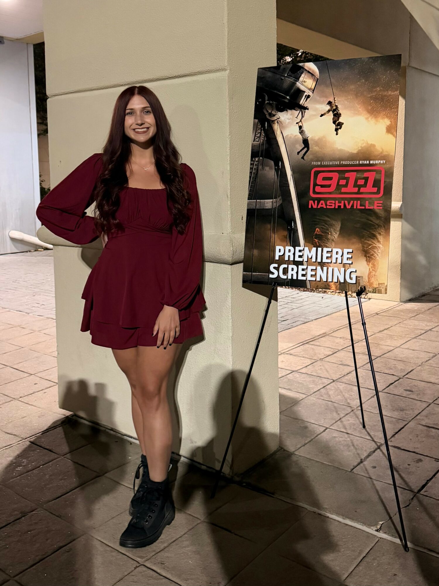 Young woman with long dark hair in a burgundy dress and black boots standing next to a poster for a TV premiere screening.