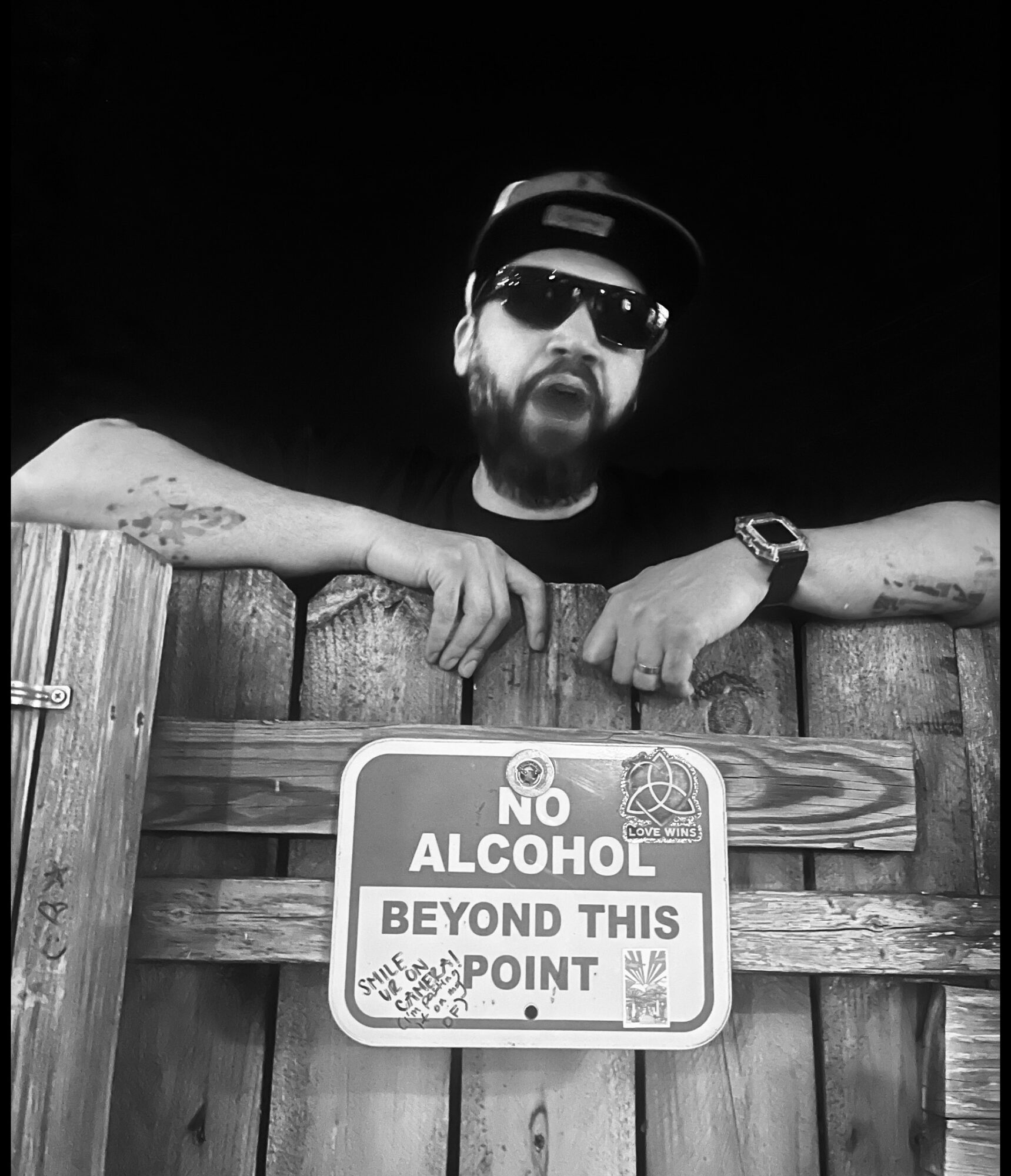 Man with sunglasses and a cap leaning on a wooden fence, holding a sign that says no alcohol beyond this point.