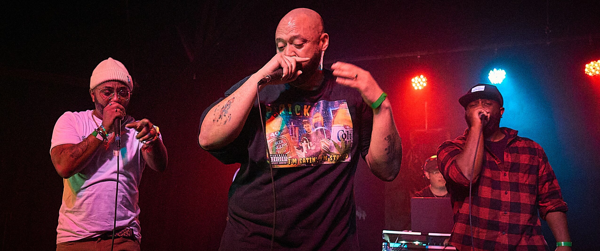 Three men perform on stage with colorful lights, one with a microphone, one adjusting his shirt, and another with a microphone.