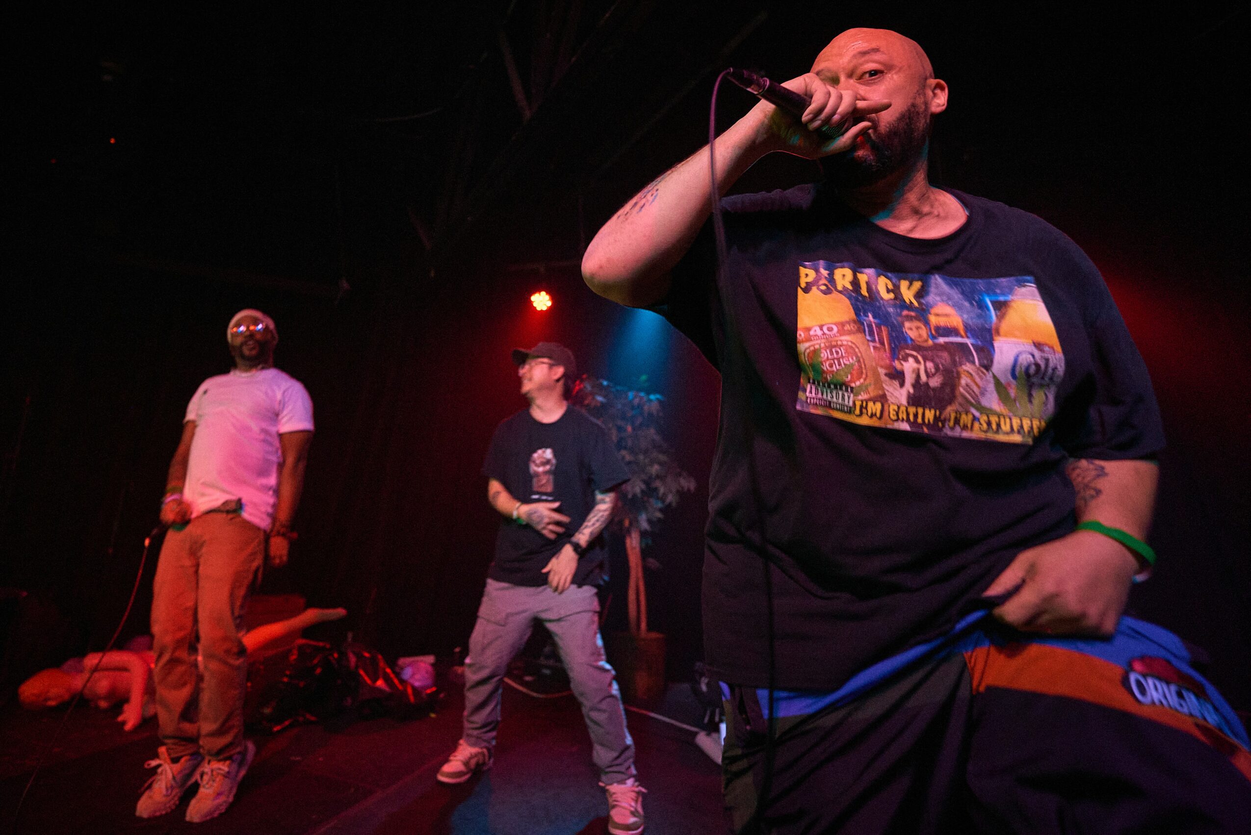 Three men on stage, one singing into a microphone, with colorful stage lighting, dark background, and equipment visible.