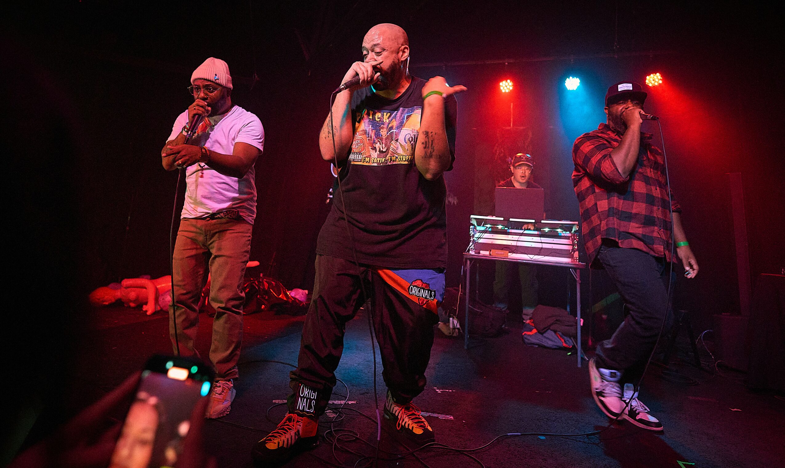 Three performers on stage with colorful lighting, one singing into a microphone, others holding microphones, DJ equipment in background.