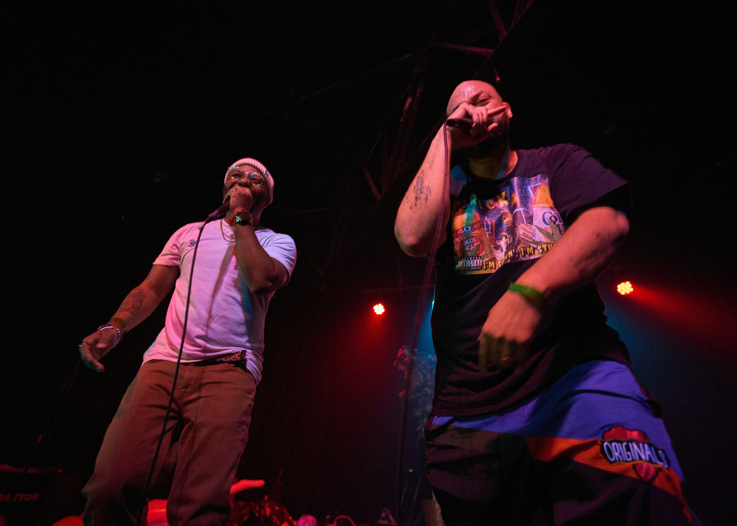 Two performers on stage, one holding a microphone, with red stage lights in the background.