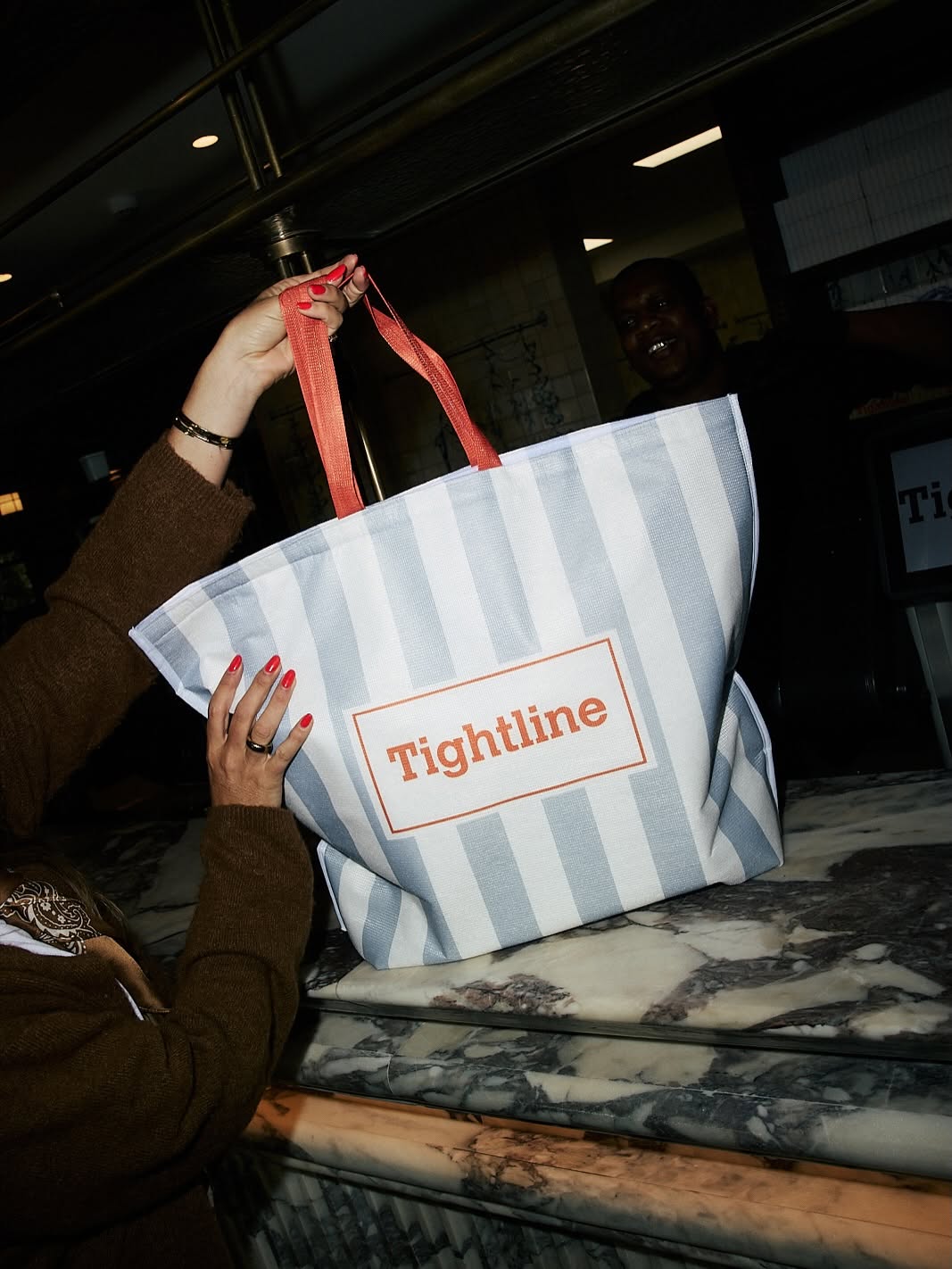 Person holding a large striped tote bag with red handles and a label reading 'Tightline' inside a store or cafe.