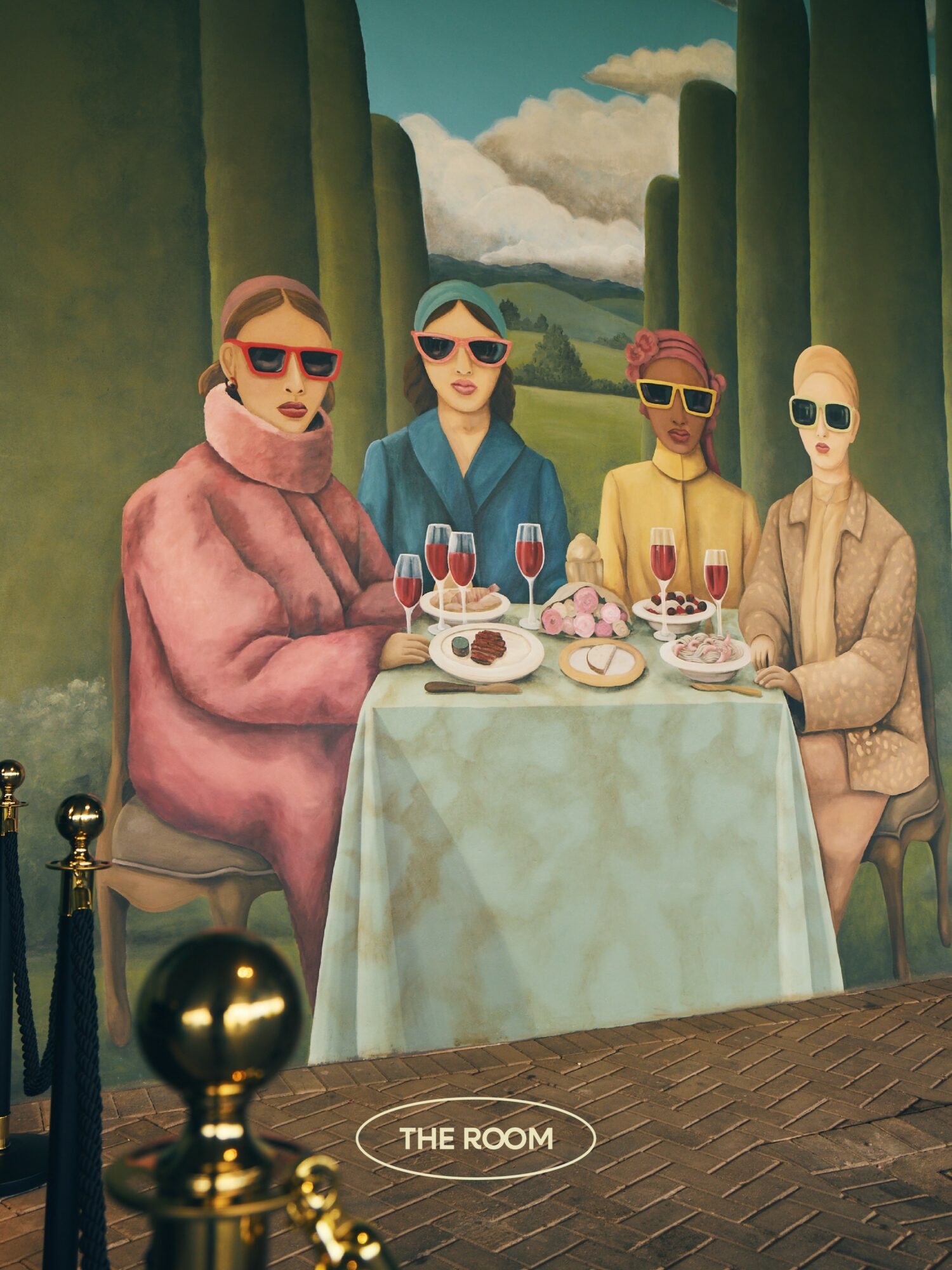 Four women sit at a table outdoors with a landscape background, wearing sunglasses and fashionable clothing, enjoying desserts and drinks.