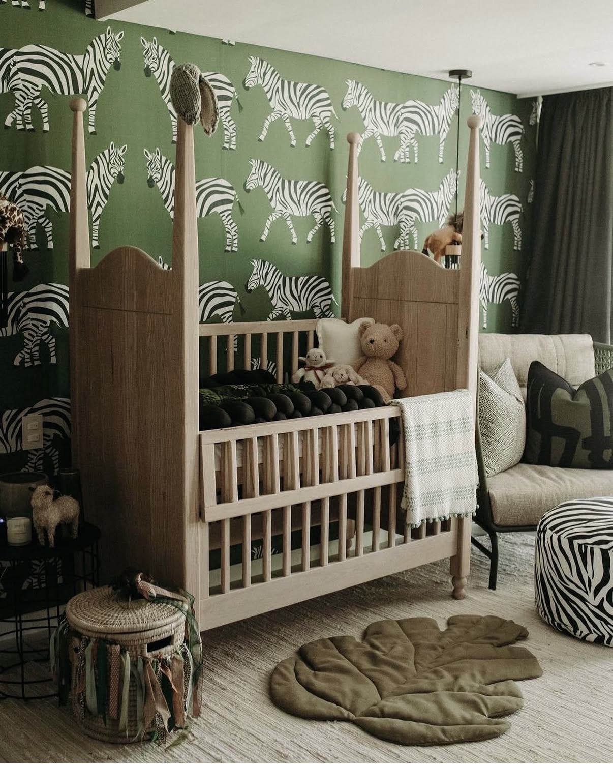 Crib with stuffed bear in a room with zebra-patterned wallpaper, a beige sofa, and a round ottoman, with a green rug on the floor.