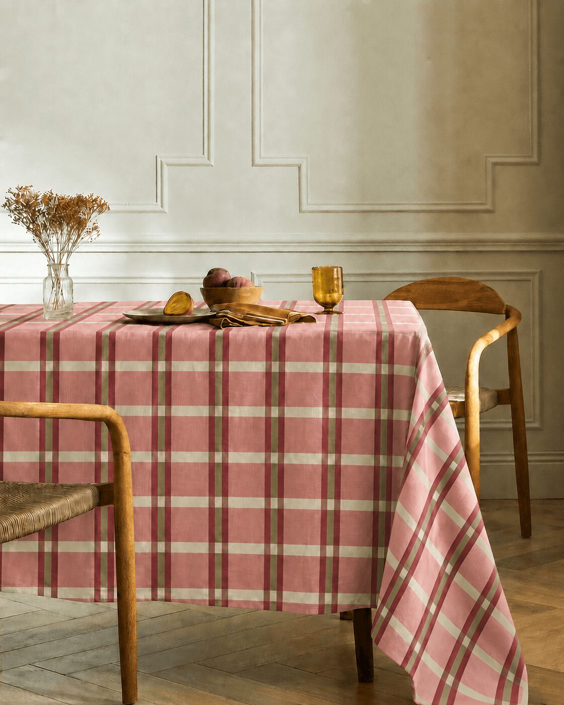 Dining table with a pink plaid tablecloth, a glass vase with dried flowers, and a yellow glass, against a white wall.