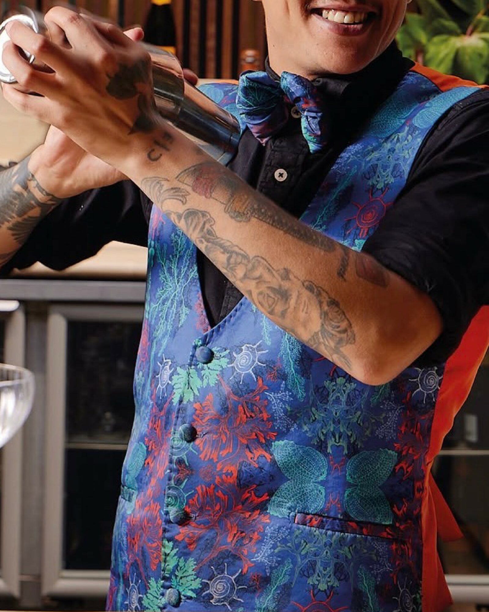 Person smiling, wearing a colorful vest and scarf, holding a cocktail shaker, with tattoos on arms, in an outdoor setting.