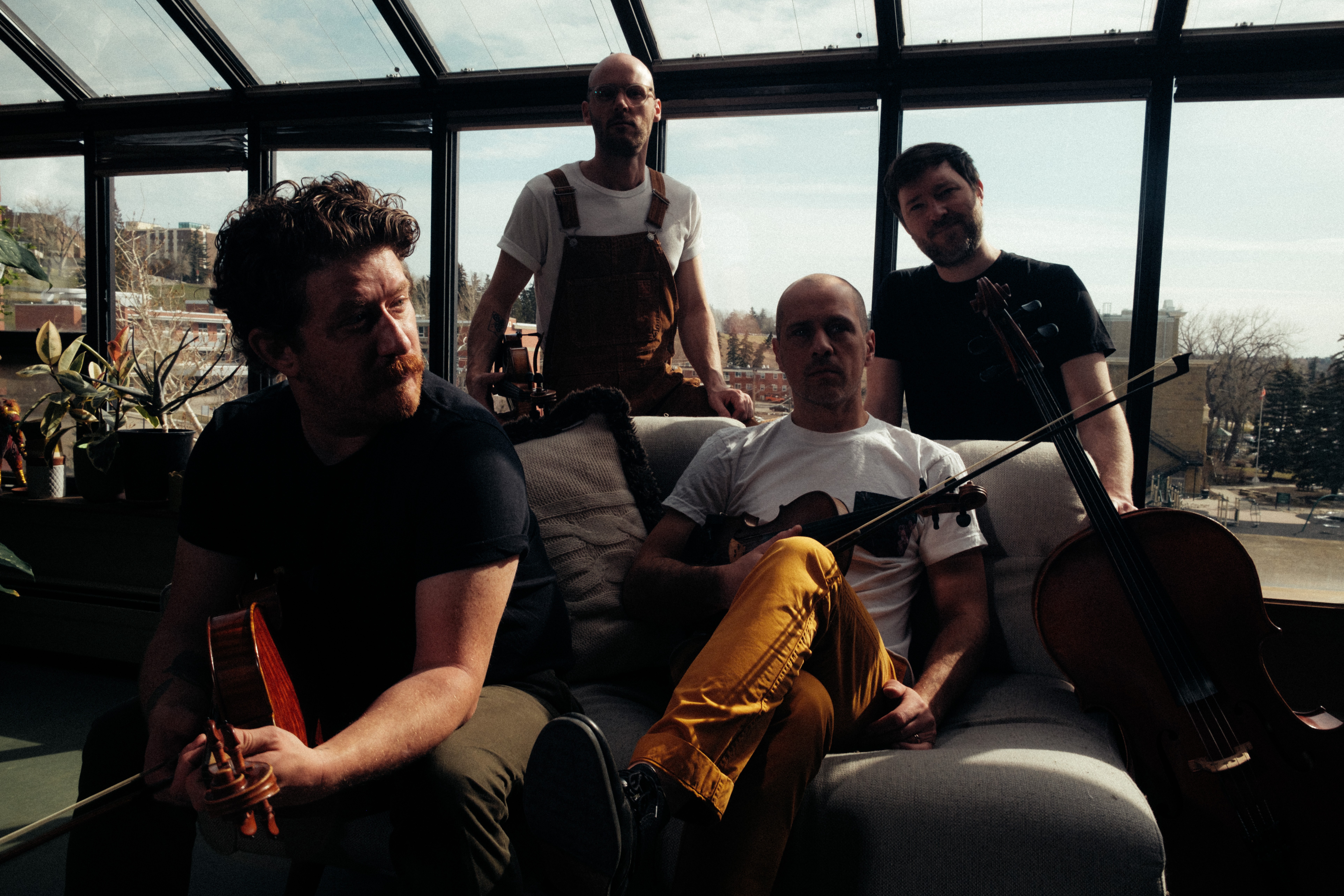 Four people sitting and standing in a sunlit room with large windows, one holding a cello, others seated on a sofa.