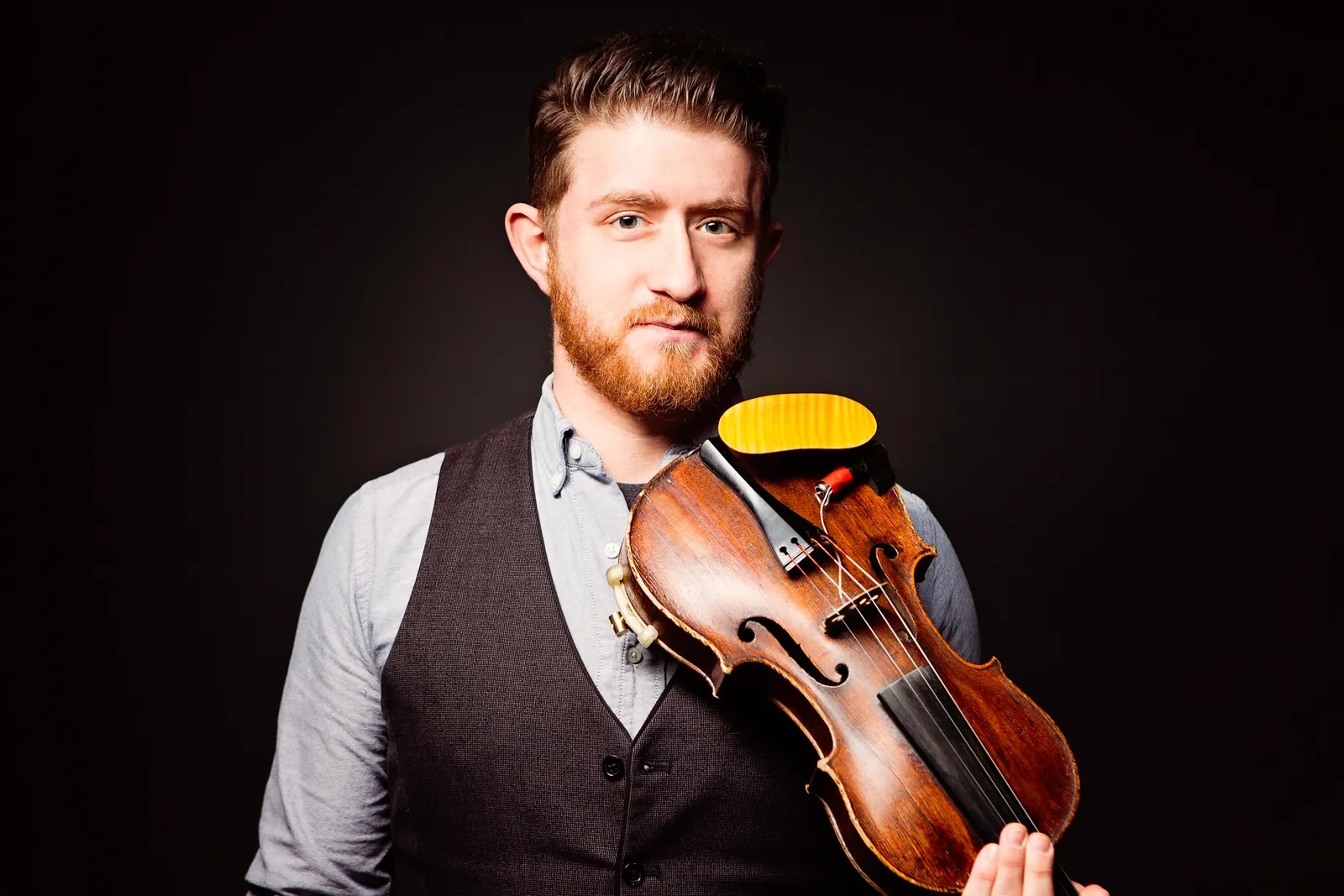 Man with a beard holding a violin against a dark background.