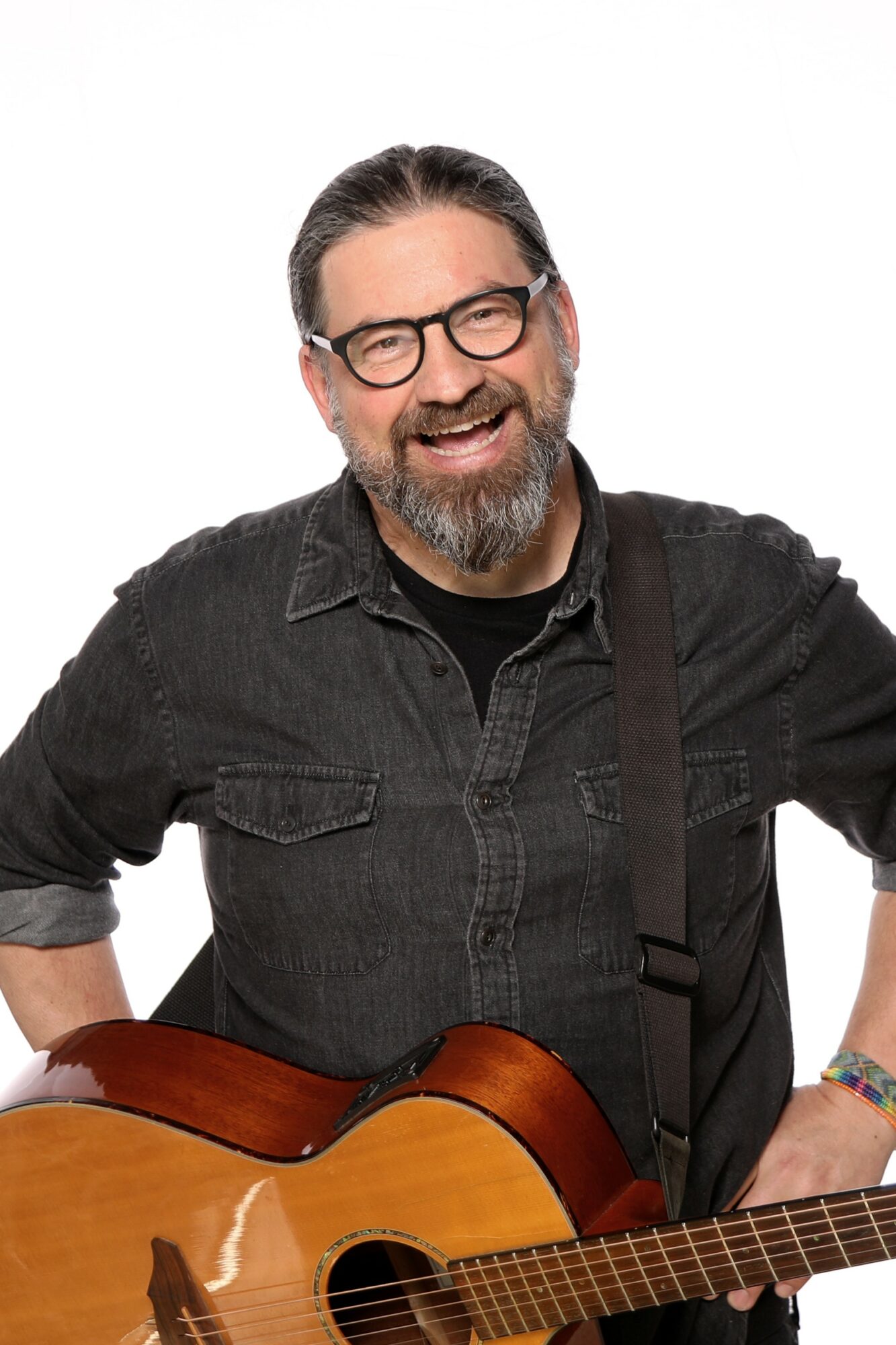Smiling man with glasses and a beard holding an acoustic guitar, wearing a black shirt, against a white background.
