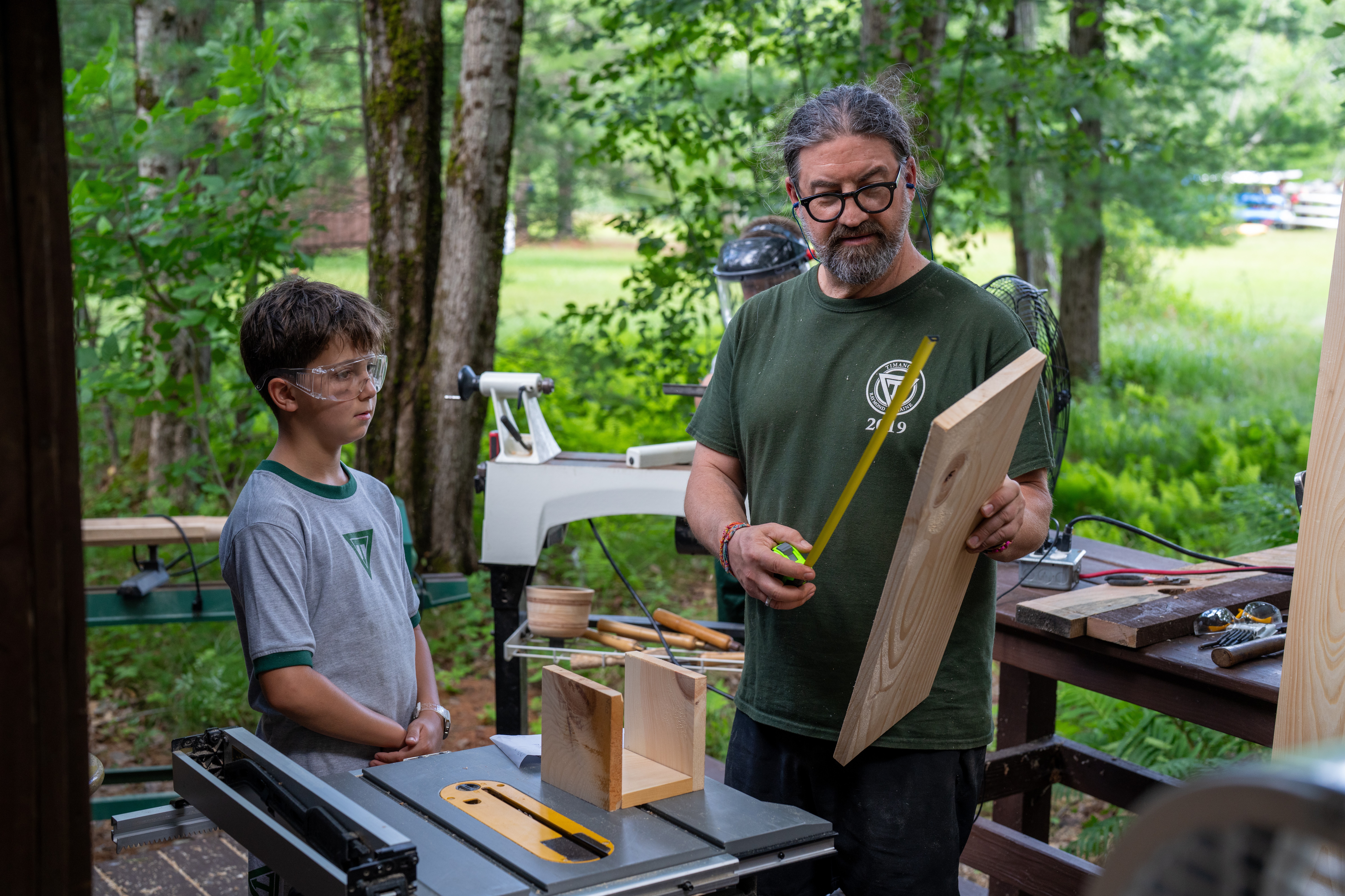 Man demonstrating woodworking to a boy outdoors with trees and greenery in background.