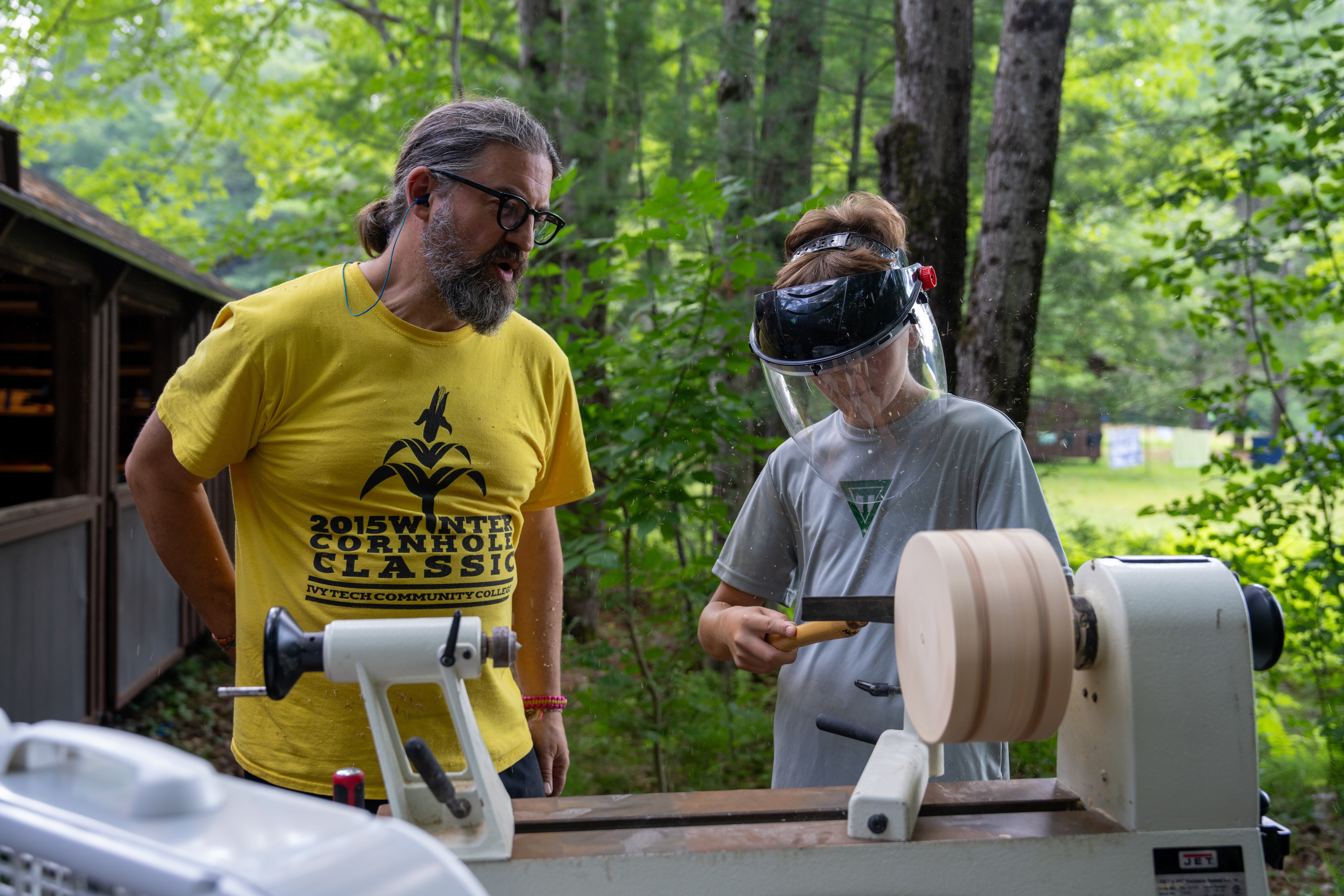 Two people outdoors in a wooded area, one demonstrating woodturning with a lathe, the other observing, both wearing safety glasses.