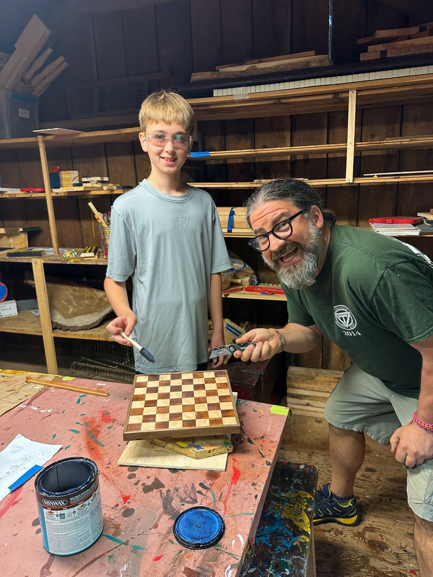 A young boy and an adult man with glasses and a beard stand in a woodworking shop, smiling near a chessboard on a table.