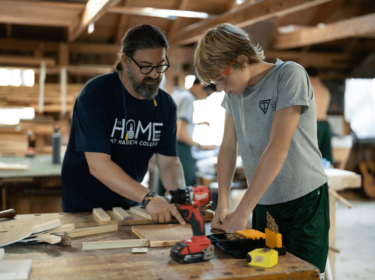 Two people working on a woodworking project in a workshop, using a power drill and measuring tools.