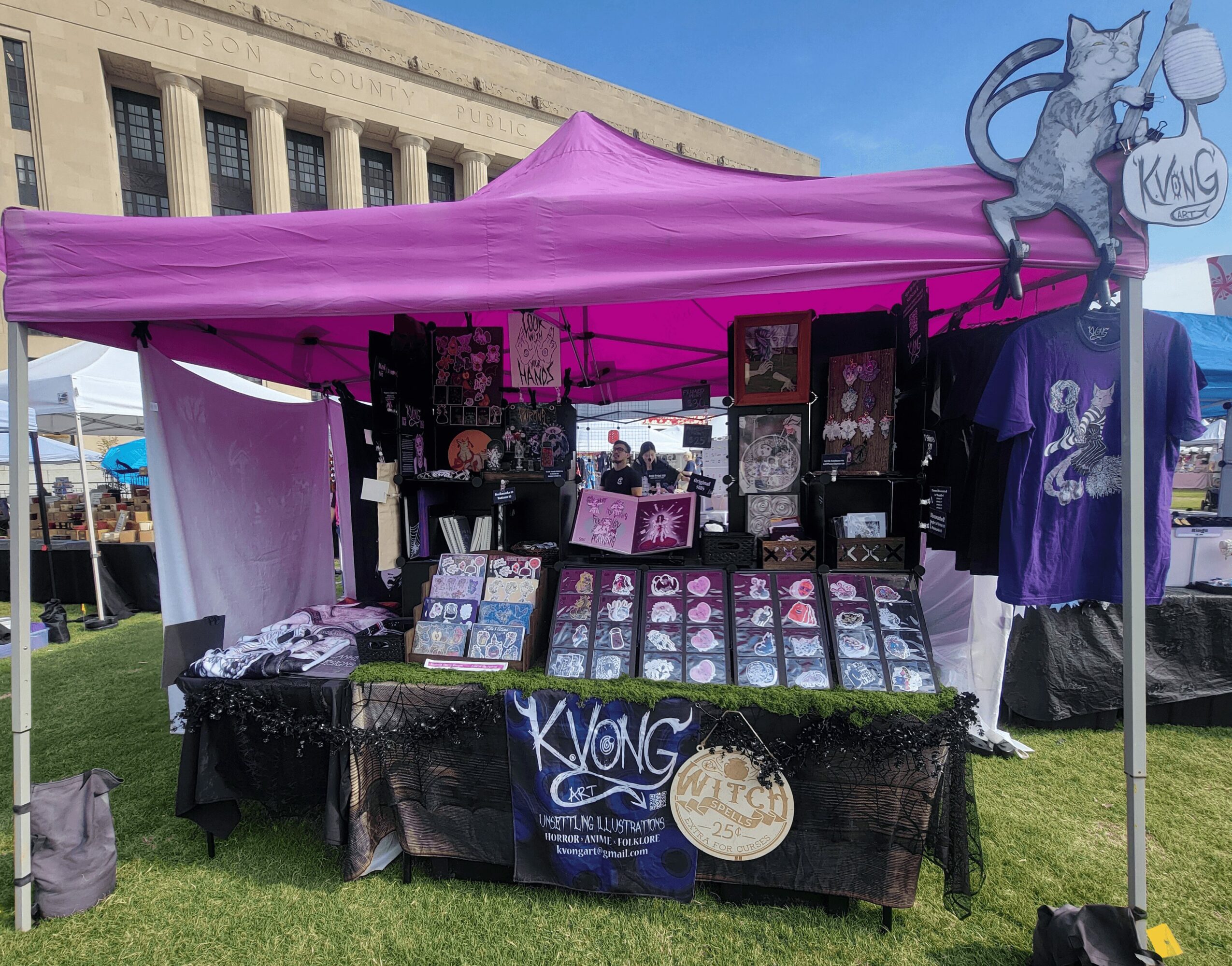 Vendors' booth with purple canopy displaying merchandise, including clothing and artwork, in an outdoor setting with a building in background.