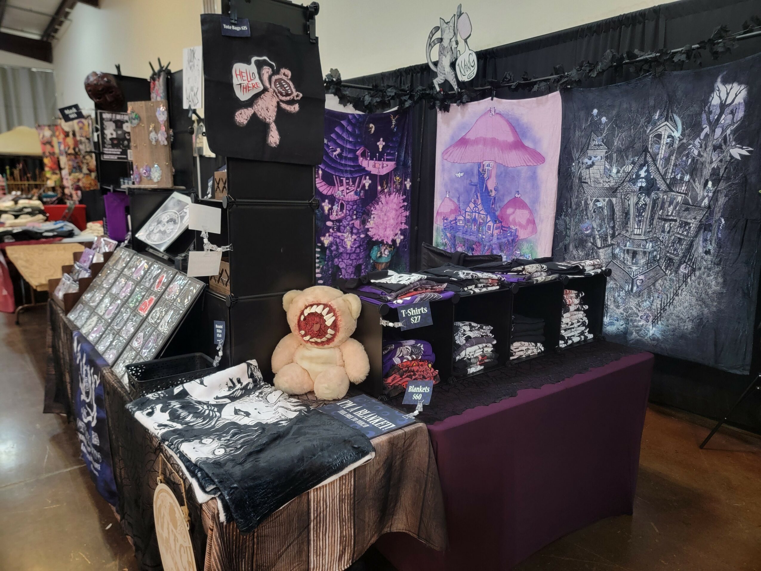 Display booth with plush teddy bear, artwork, and decorative items on tables and hanging fabric backdrop.