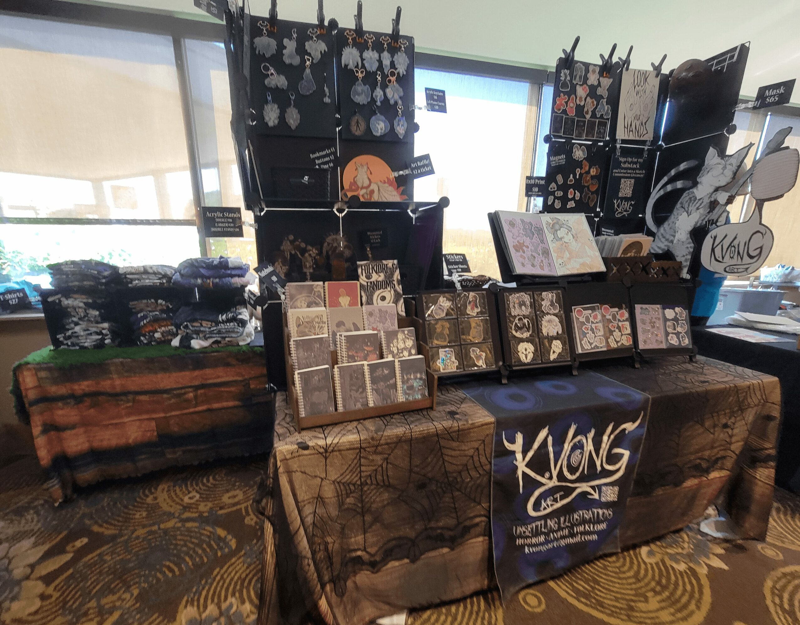 Display table with jewelry, artwork, and accessories at a craft fair, with hanging earrings and framed art pieces.