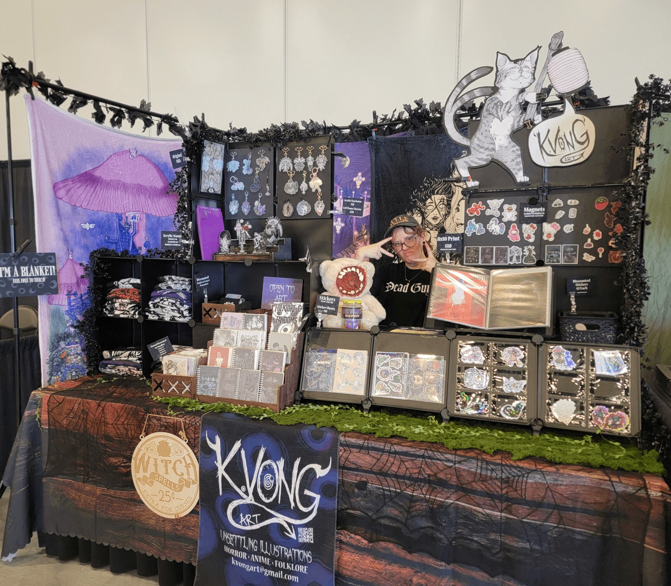 Vendor booth displaying jewelry, artwork, and accessories with a person behind the counter, decorated with black garland and a large cat figure. Word 'KVONG' visible.