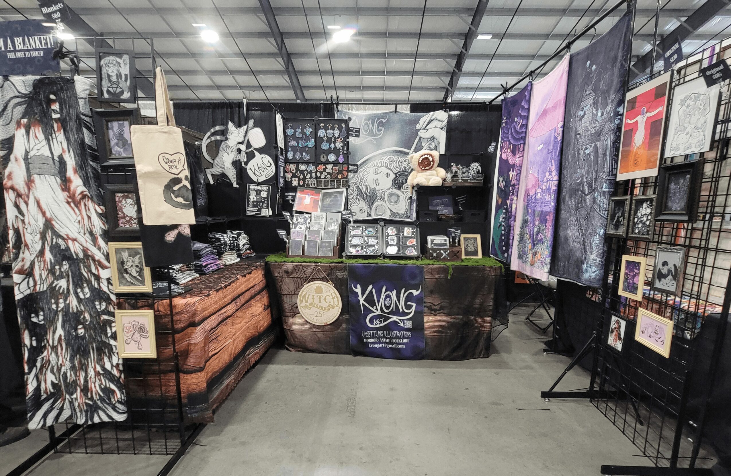 Art and craft booth with paintings, prints, and decorative items displayed on tables and grid panels inside a large indoor space.