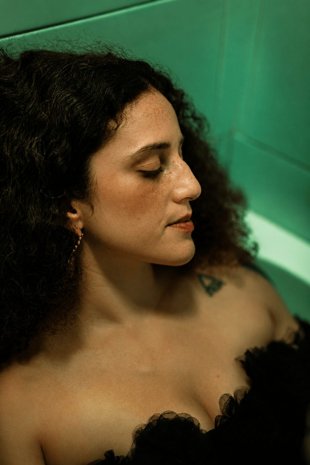 Side profile of a woman with curly hair and earrings, eyes closed, against a green background.