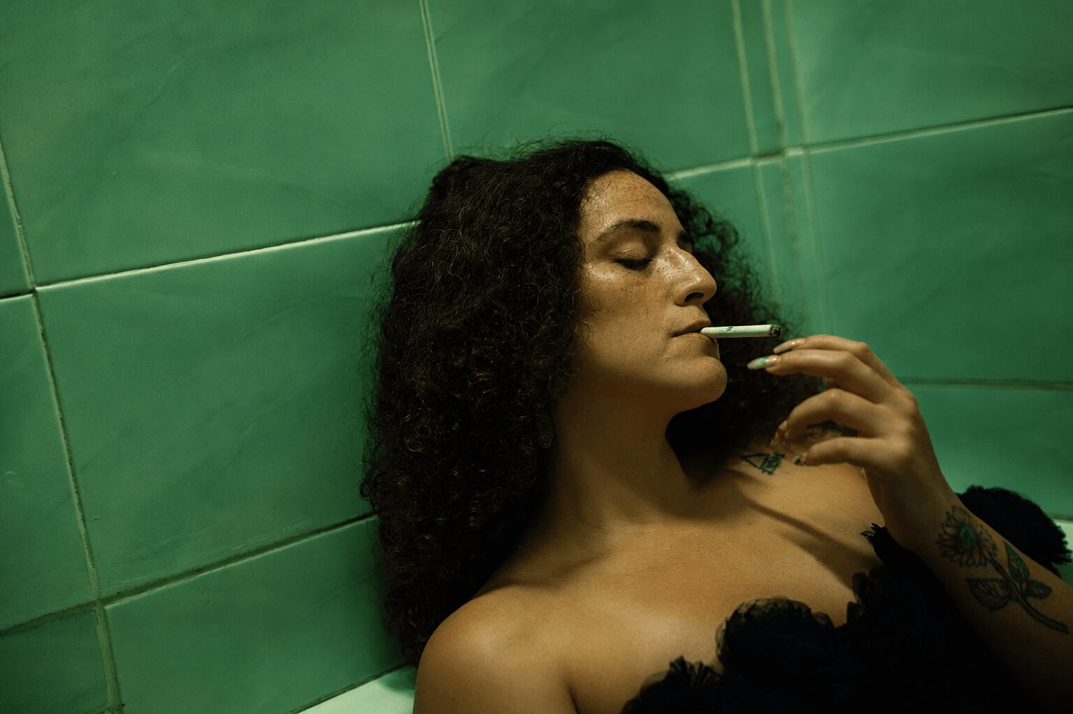 Woman with curly hair leaning against green tiled wall, smoking a cigarette, with a tattoo on her arm.