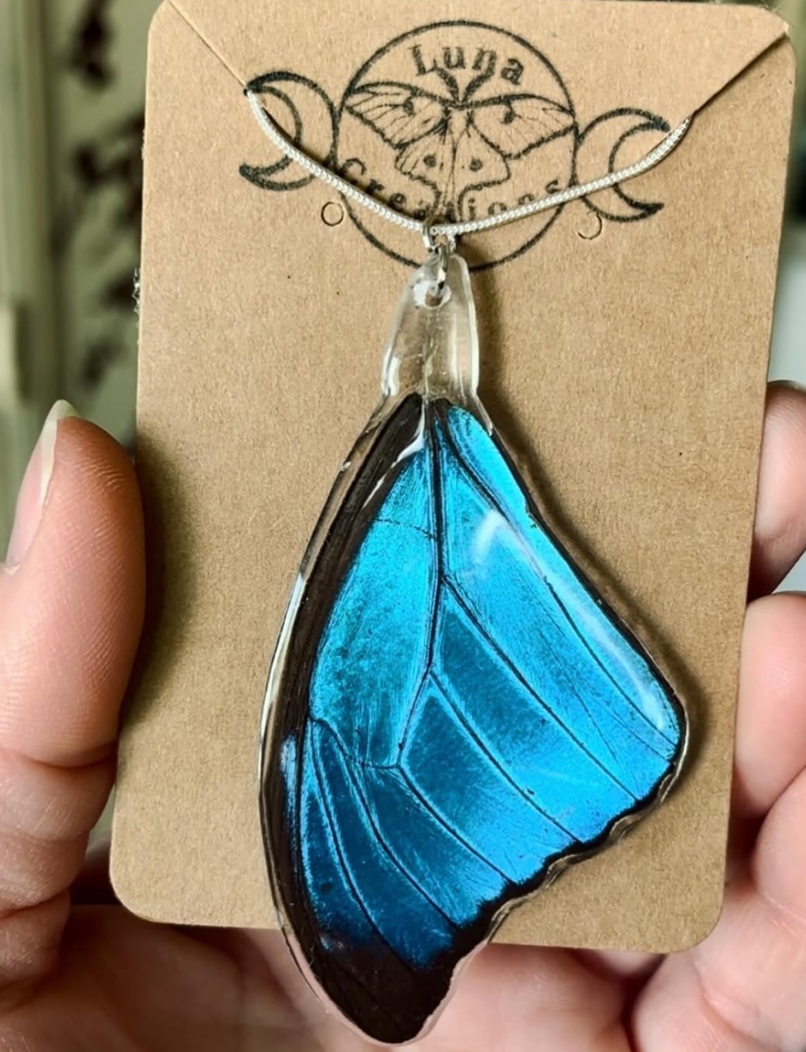Blue butterfly-shaped pendant necklace on cardboard backing with logo and text, held by a person's hand.