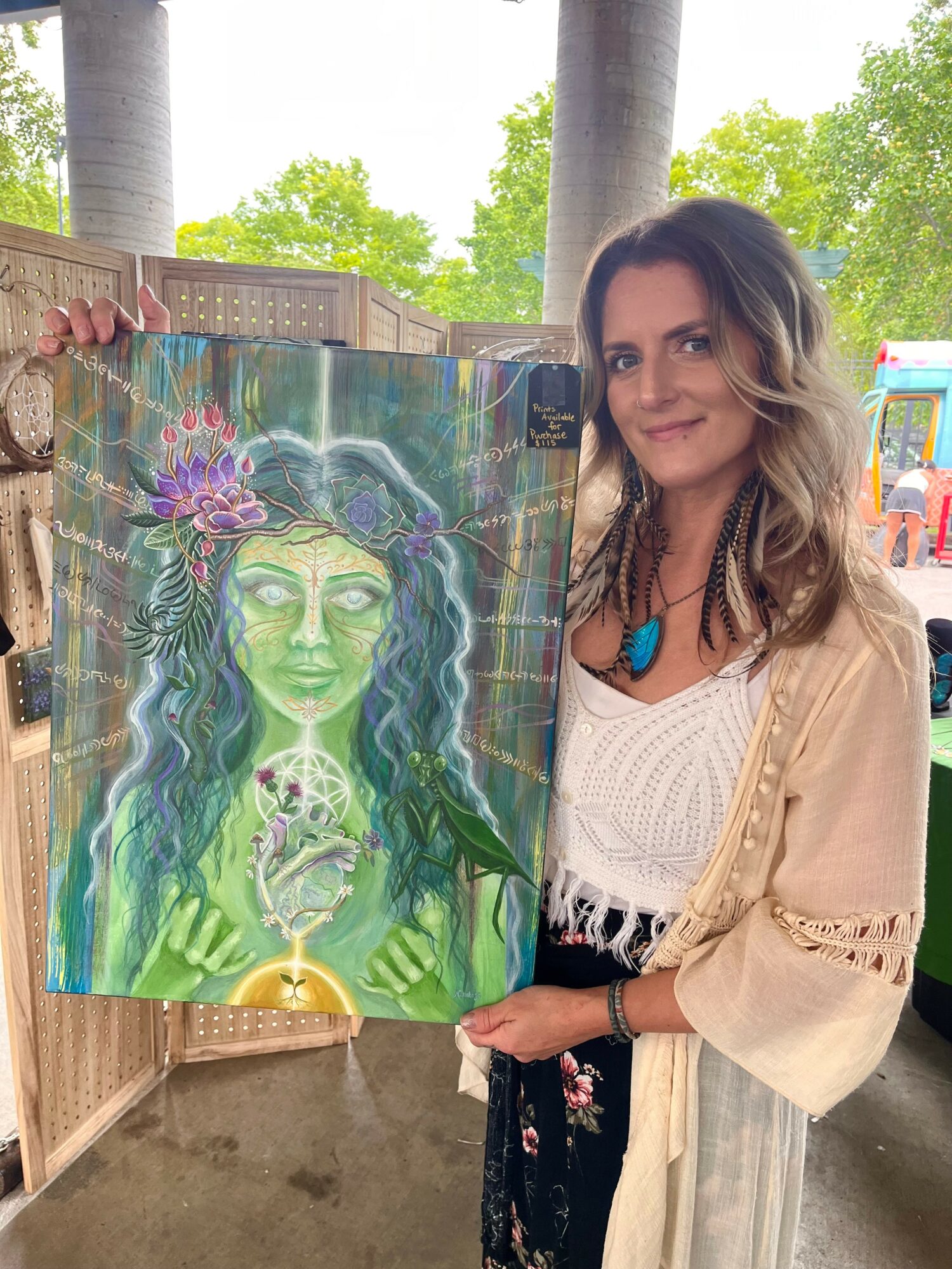 Woman with long wavy hair holding a colorful painting of a green-skinned woman with flowers and a snake, outdoors.
