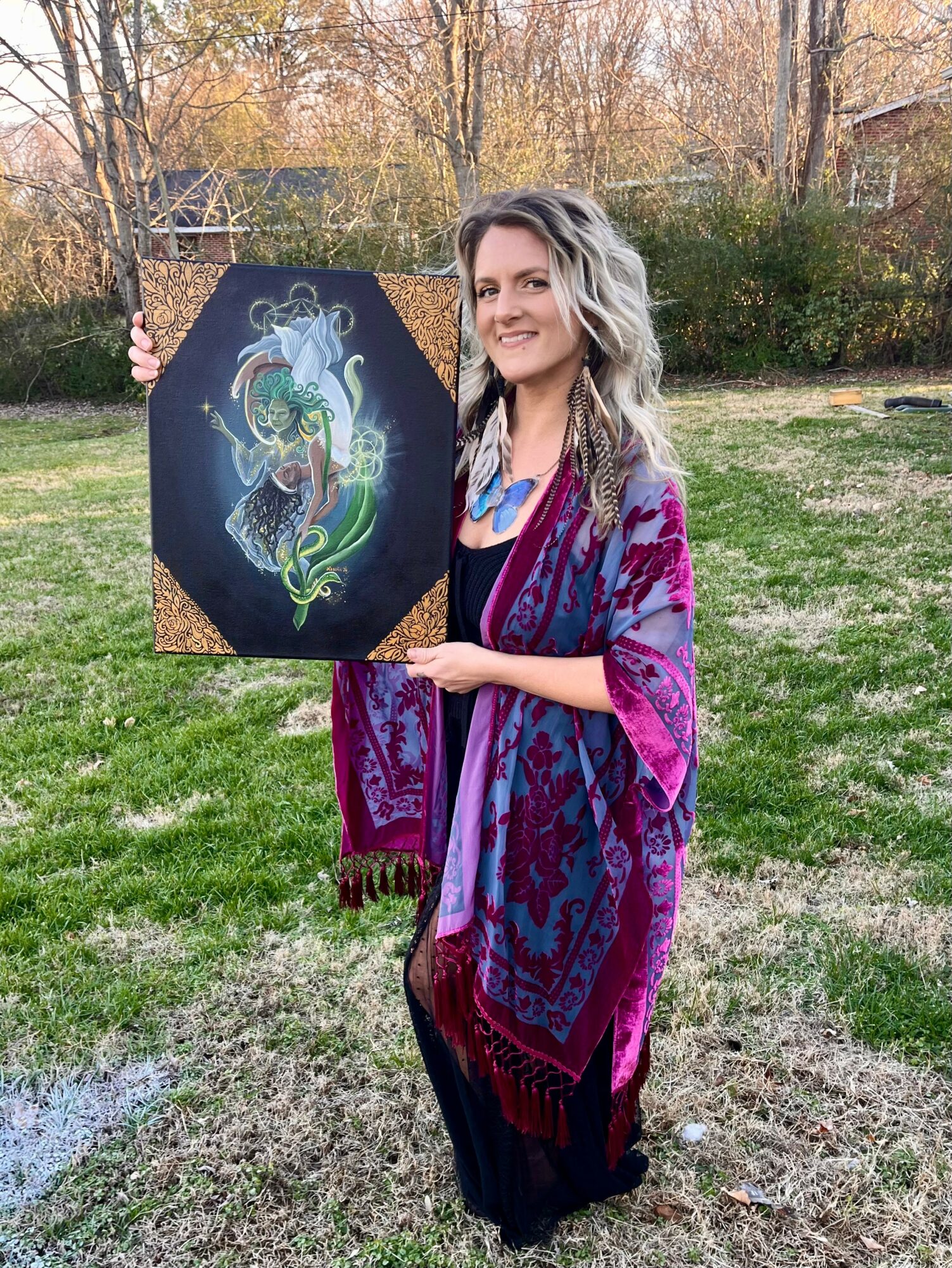 Woman with long wavy hair holding a colorful dragon artwork outdoors, wearing a purple and blue patterned shawl, smiling.