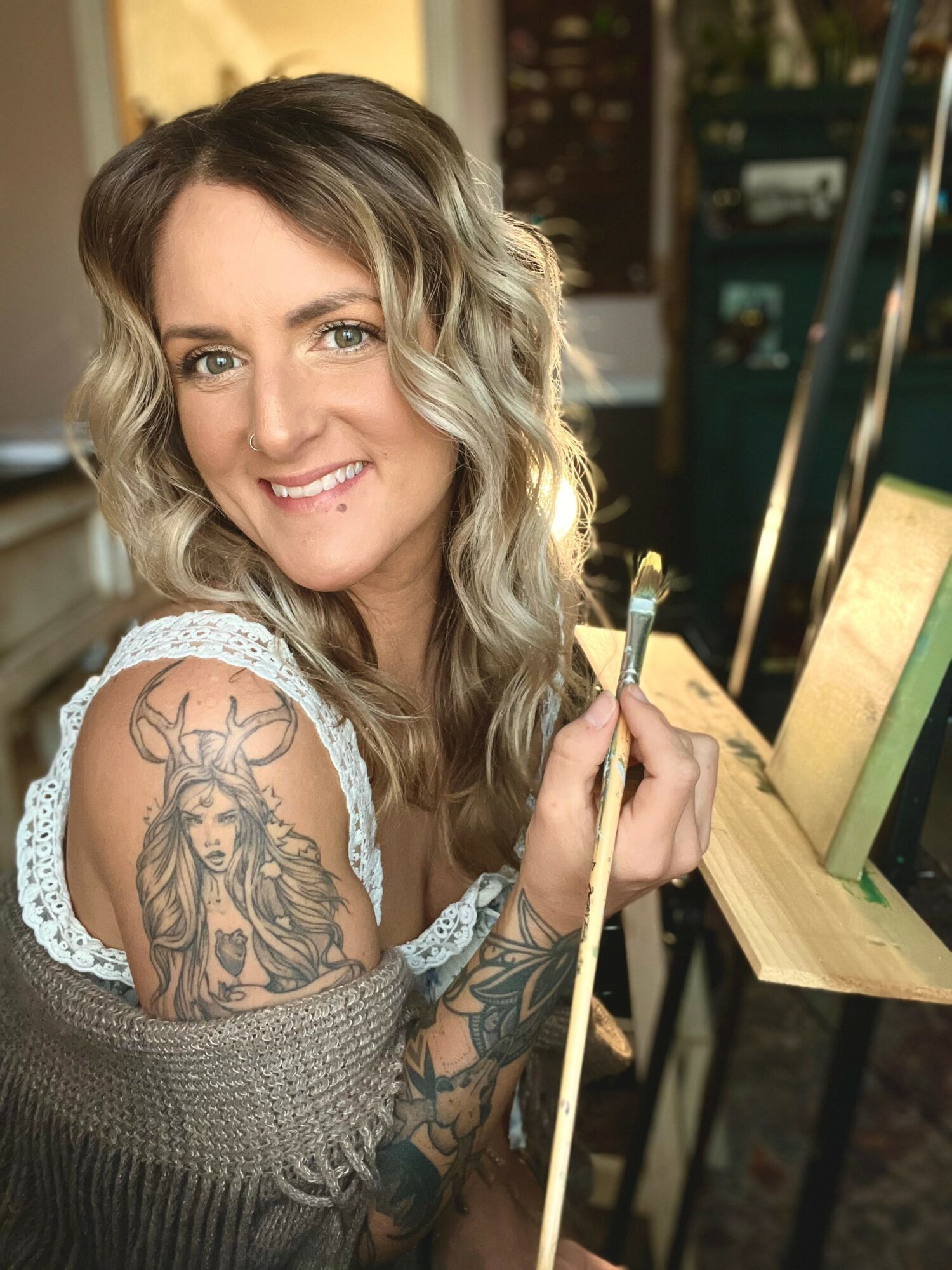Woman with wavy hair smiling, holding a pool cue, sitting at a gaming table, tattoo of a deer on her arm, indoor setting.