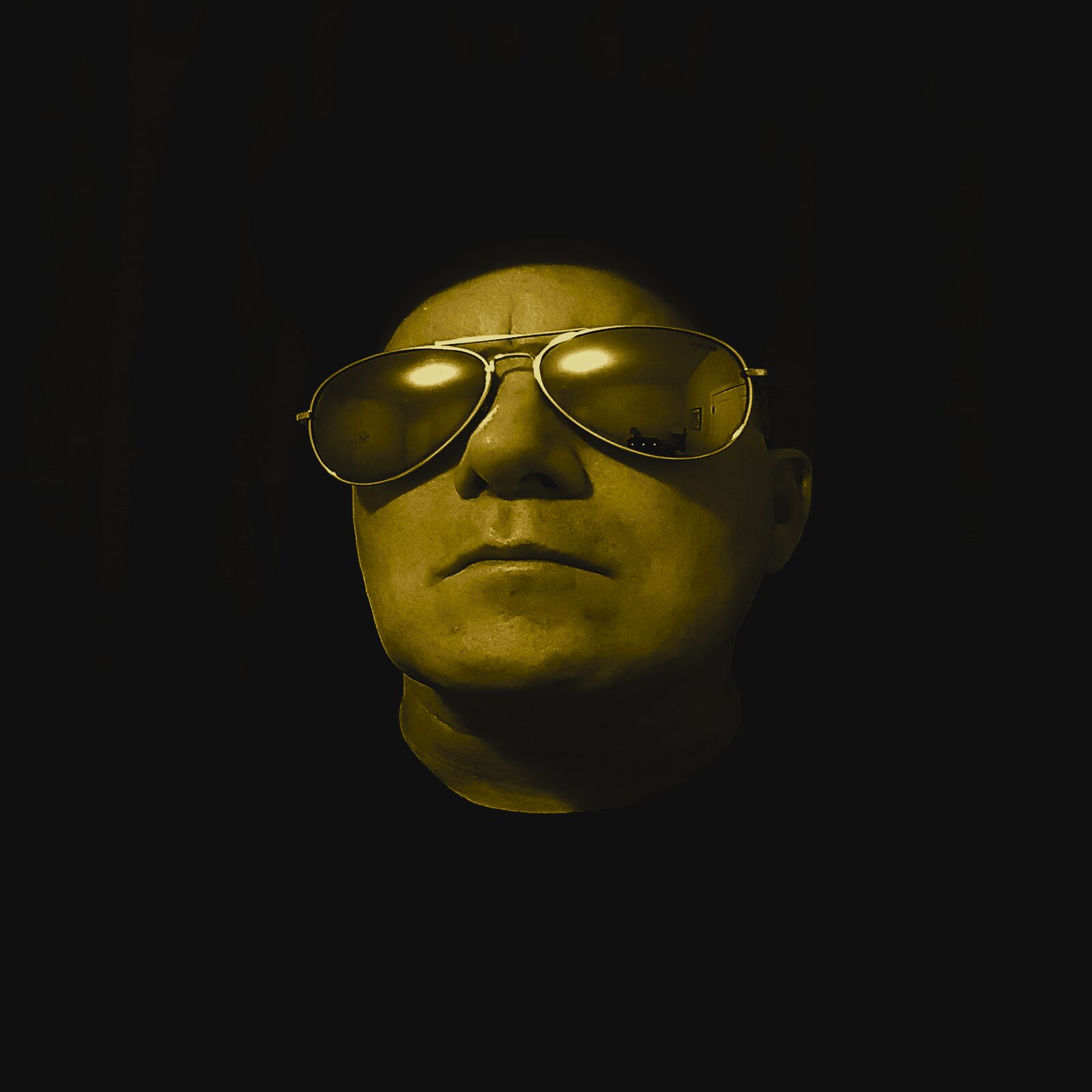 Person wearing sunglasses with a serious expression, head slightly tilted, dark background, yellowish lighting.