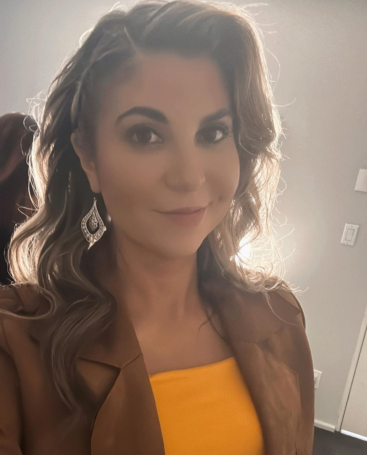 Woman with wavy hair, wearing earrings and a yellow top, smiling at the camera.