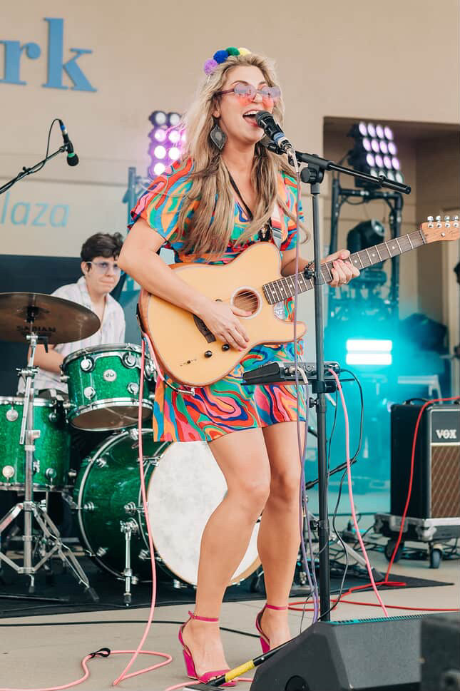 Woman singing and playing guitar on stage with drummer in background, colorful dress, glasses, and pink shoes.