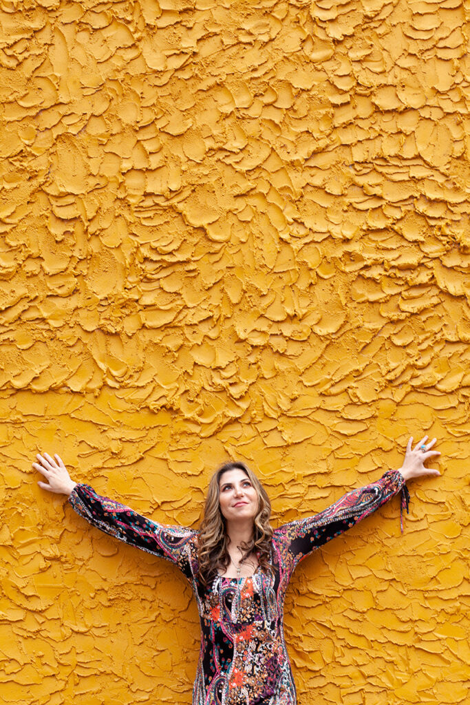 Woman with arms outstretched standing in front of a textured yellow wall.