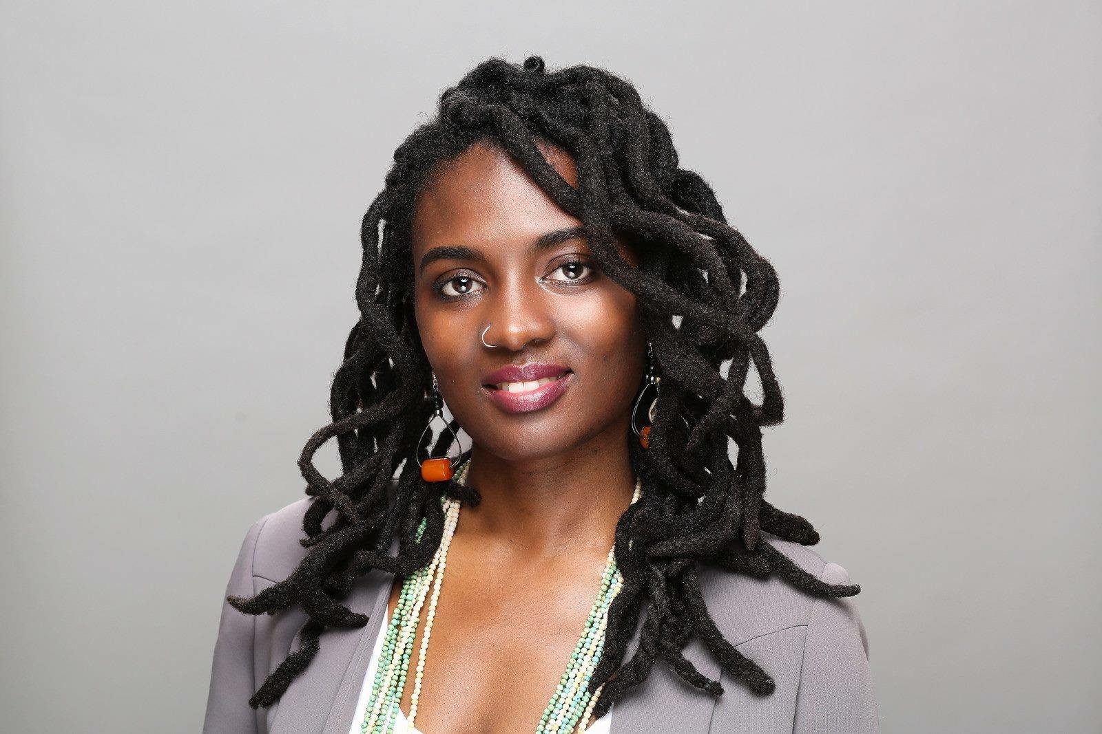 Portrait of a woman with long, curly hair wearing a light-colored blazer and earrings, smiling at the camera.