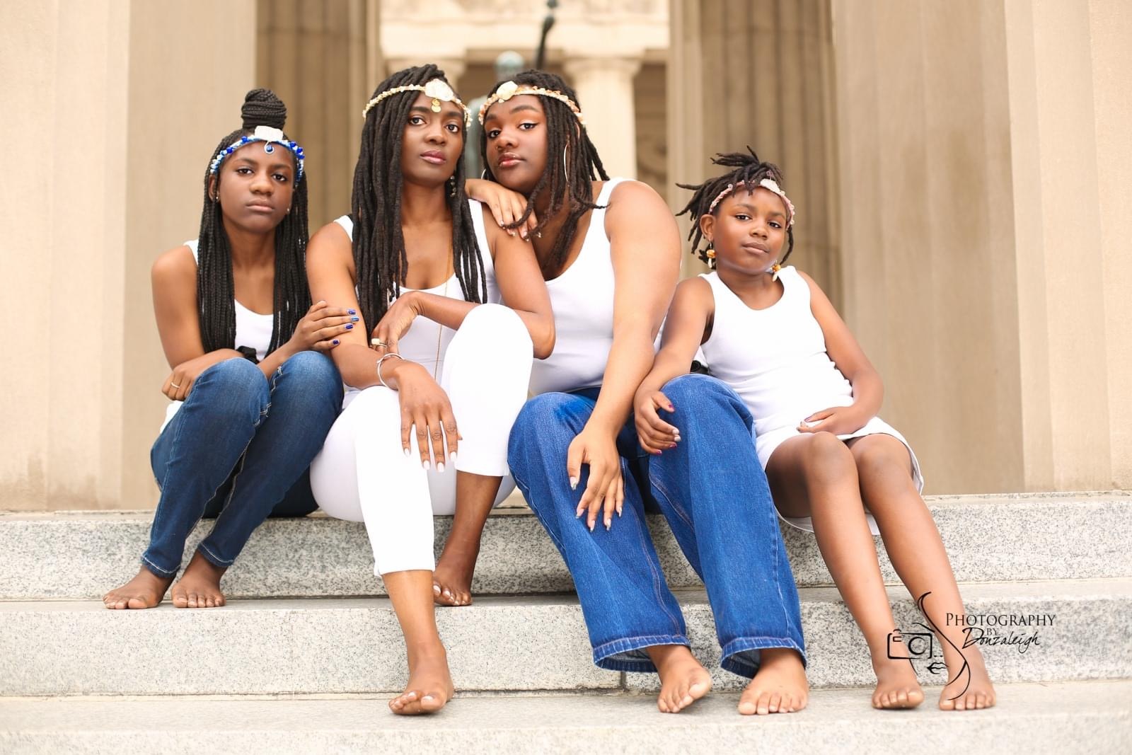 Four young girls sit on steps in front of a building with columns, wearing casual clothing and headbands.